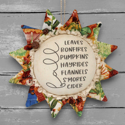 A fabric sunflower-shaped ornament with a rustic autumn theme, featuring a central cream-colored circle with black text listing fall foods and flavors, surrounded by patterned fabric points in orange, green, blue, and red tones, hanging against a gray wood plank background.