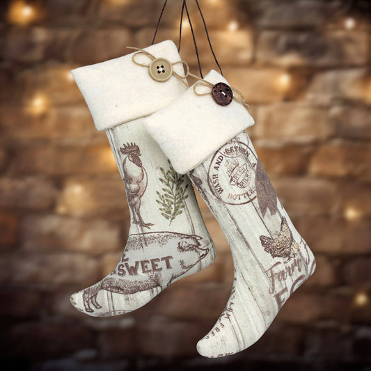 Two rustic Christmas stockings hanging from black strings against a blurred warm brick wall with bokeh lights. The stockings are off-white with vintage-style brown printed graphics including chickens, a rooster, a dog, and text such as "SWEET" and "Farm.