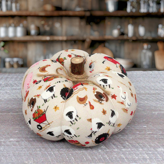A fabric-covered decorative pumpkin with a rustic stem, featuring a printed pattern of sheep, chickens, stars, and a red wheelbarrow on an off-white background, displayed on a gray textured surface with a blurred rustic shop background.