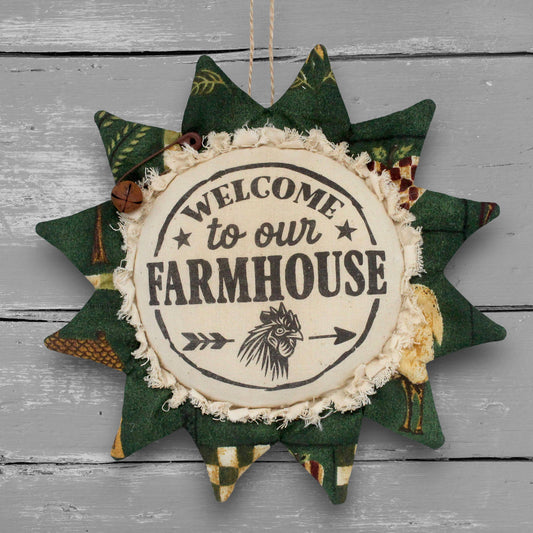 A fabric sunflower-shaped hanging ornament with a rustic farmhouse design. The center is cream-colored fabric with the words 'WELCOME to our FARMHOUSE' and a rooster illustration, surrounded by green patterned starburst points and a small wooden bell accent, hanging against a gray wood plank background.
