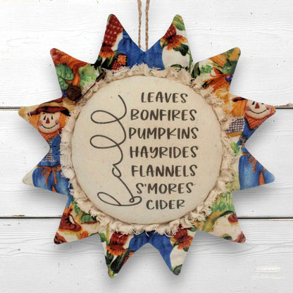 A fabric sunflower-shaped hanging ornament with a rustic cream center that reads 'LEAVES BONFIRES PUMPKINS HAYRIDES FLANNELS S'MORES CIDER' in black script and block lettering. The sun's rays feature colorful autumn-themed fabric prints, including scarecrows, pumpkins, and fall foliage, and it hangs from a twine string against a white wood plank background.