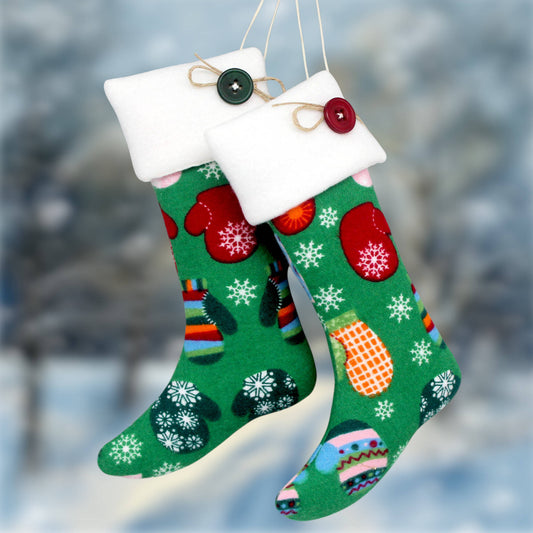 Two green Christmas stocking ornaments with white fleece cuffs and decorative buttons, hanging against a blurred snowy outdoor background.