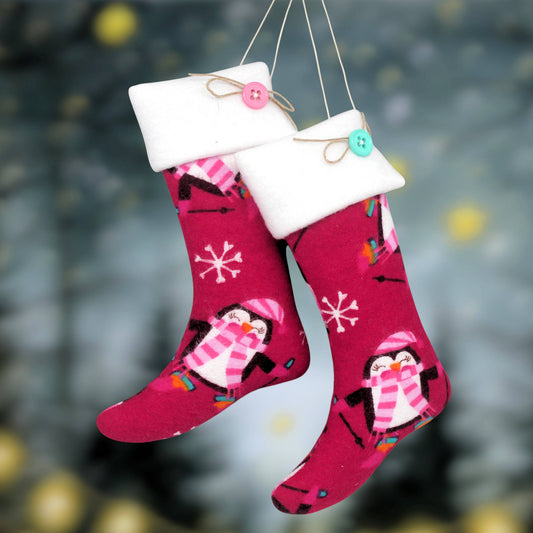 Two festive Christmas stockings hanging from strings against a blurred holiday background. The stockings are dark pink with white fleece trim, decorated with penguin illustrations wearing pink and white striped scarves, and accented with small colorful details.