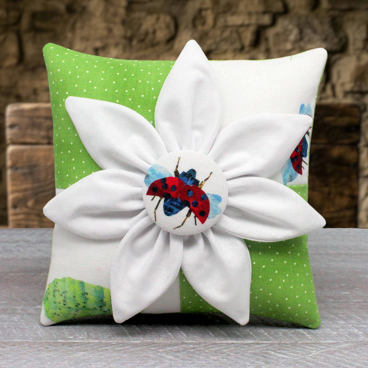 A square decorative miniature pillow with a large fabric flower on the front. The pillow has a green and white patterned fabric base with a ladybug motif, and the flower features white petals with a red and blue ladybug button in the center.