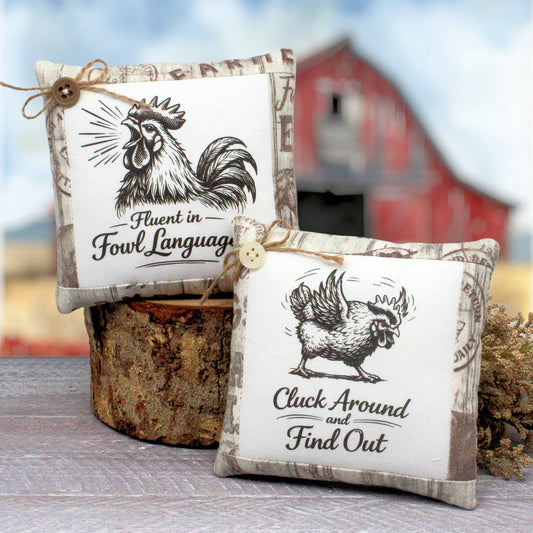 Two decorative mini pillows with rustic farmhouse-style designs, featuring black rooster illustrations and humorous text on a white center panel with beige patterned borders. One pillow is tied with twine and button, and both are styled on a rustic wood stump with a blurred red barn and blue sky background.