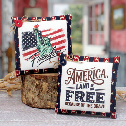 Two patriotic fabric mini pillows displayed on a rustic wood slice stand. One pillow features a distressed American flag, the Statue of Liberty, and the word 'Freedom' with a red button accent.