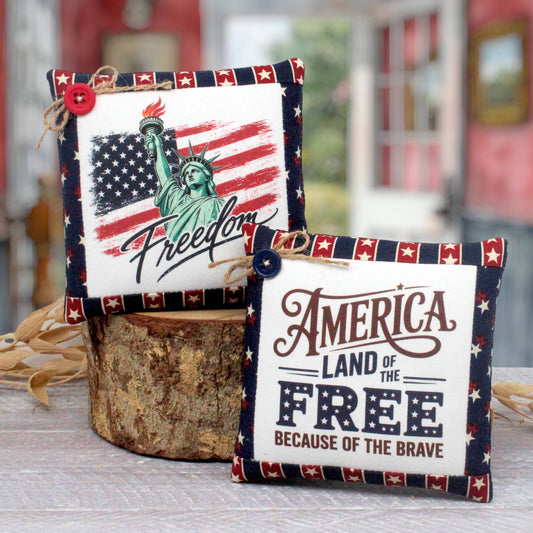 Two patriotic fabric mini pillows displayed on a rustic wood slice stand. One pillow features a distressed American flag, the Statue of Liberty, and the word 'Freedom' with a red button accent.