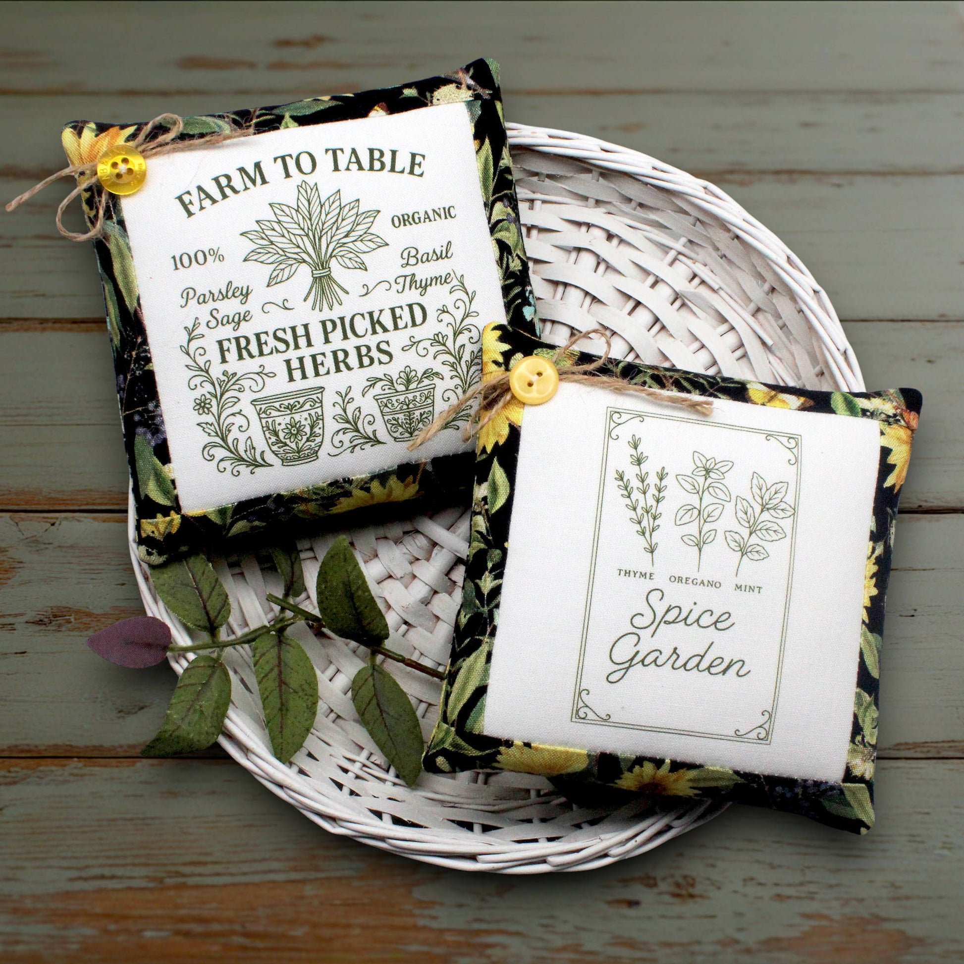 Two decorative square fabric mini pillows with floral borders and yellow button closures, placed on a white woven tray over a rustic painted wood surface. One sachet reads “Farm to Table 100% Organic Parsley Sage Basil Thyme Fresh Picked Herbs,” and the other reads “Spice Garden” with botanical illustrations.