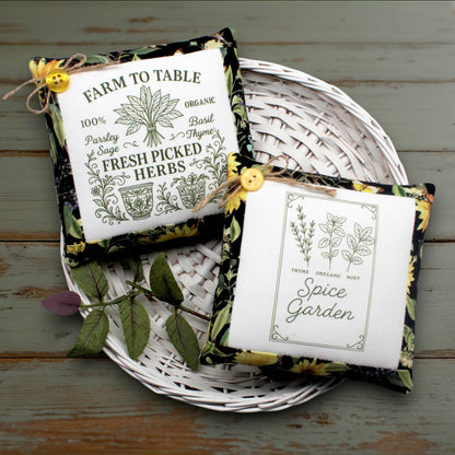 Two decorative square fabric mini pillows with floral borders and yellow button closures, placed on a white woven tray over a rustic painted wood surface. One sachet reads “Farm to Table 100% Organic Parsley Sage Basil Thyme Fresh Picked Herbs,” and the other reads “Spice Garden” with botanical illustrations.