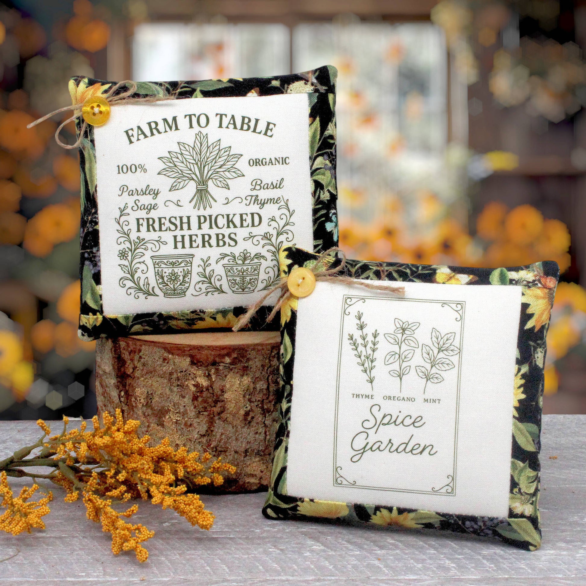 Two decorative square fabric mini pillows displayed on a rustic wood block with autumn flowers in the foreground. One pillow reads 'Farm to Table' and the other reads 'Spice Garden,' both with floral borders and yellow buttons.