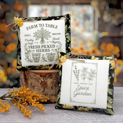 Two decorative square fabric mini pillows displayed on a rustic wood block with autumn flowers in the foreground. One pillow reads 'Farm to Table' and the other reads 'Spice Garden,' both with floral borders and yellow buttons.