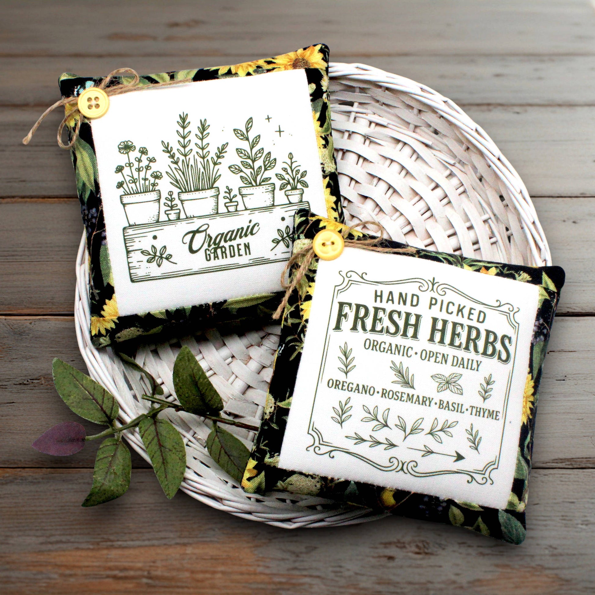 Two square fabric herb mini pillows with printed designs, displayed on a white woven tray over a rustic wooden surface. One label reads 'Organic Garden' with plant illustrations, and the other reads 'Hand Picked Fresh Herbs' with herb illustrations.