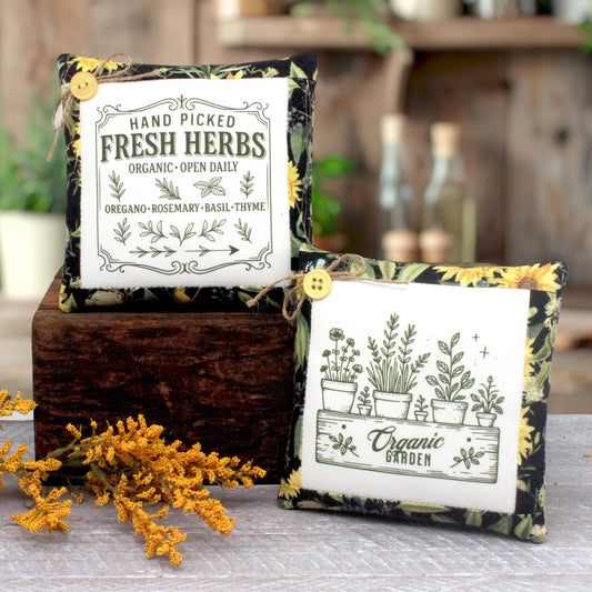 Two decorative fabric herb mini pillows with printed botanical designs, displayed on a rustic wooden block outdoors. One sachet reads 'Fresh Herbs' and the other 'Organic Garden,' both with yellow sunflower and green leaf borders, yellow buttons, and twine.