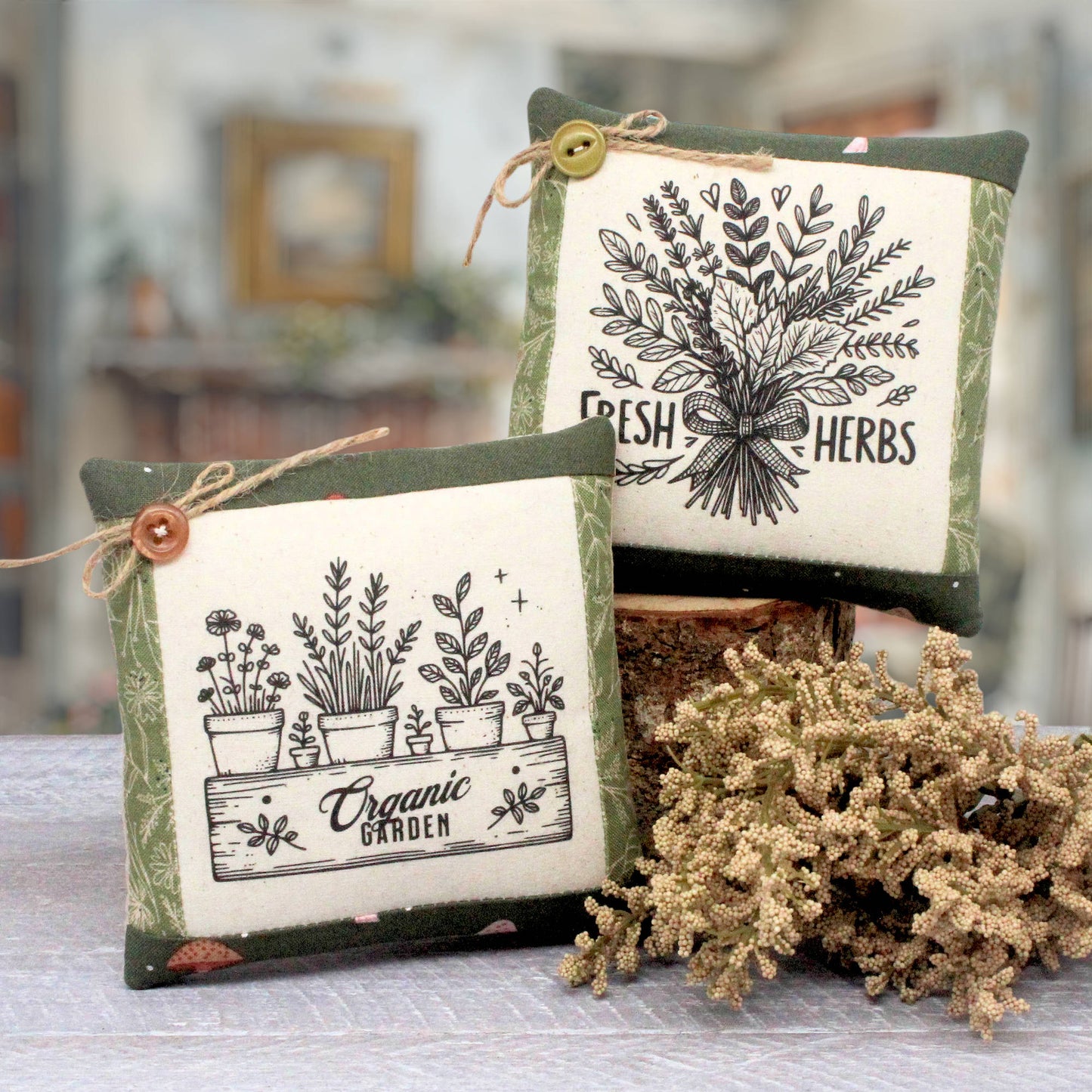 Two decorative square throw pillows with a rustic botanical theme, displayed on a table with dried flowers. One pillow features a black and white illustration of potted herbs labeled 'Organic Garden' with a brown button and twine, and the other features a black and white illustration of fresh herbs labeled 'Fresh Herbs' with a yellow button and twine.