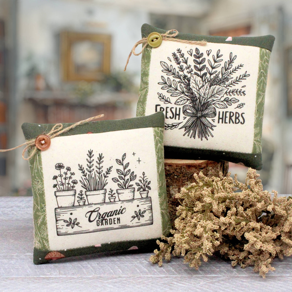 Two decorative square throw pillows with a rustic botanical theme, displayed on a table with dried flowers. One pillow features a black and white illustration of potted herbs labeled 'Organic Garden' with a brown button and twine, and the other features a black and white illustration of fresh herbs labeled 'Fresh Herbs' with a yellow button and twine.