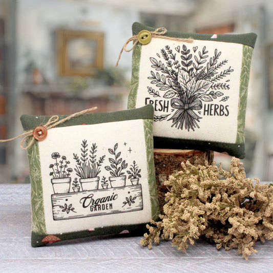 Two decorative square throw pillows with a rustic botanical theme, displayed on a table with dried flowers. One pillow features a black and white illustration of potted herbs labeled 'Organic Garden' with a brown button and twine, and the other features a black and white illustration of fresh herbs labeled 'Fresh Herbs' with a yellow button and twine.