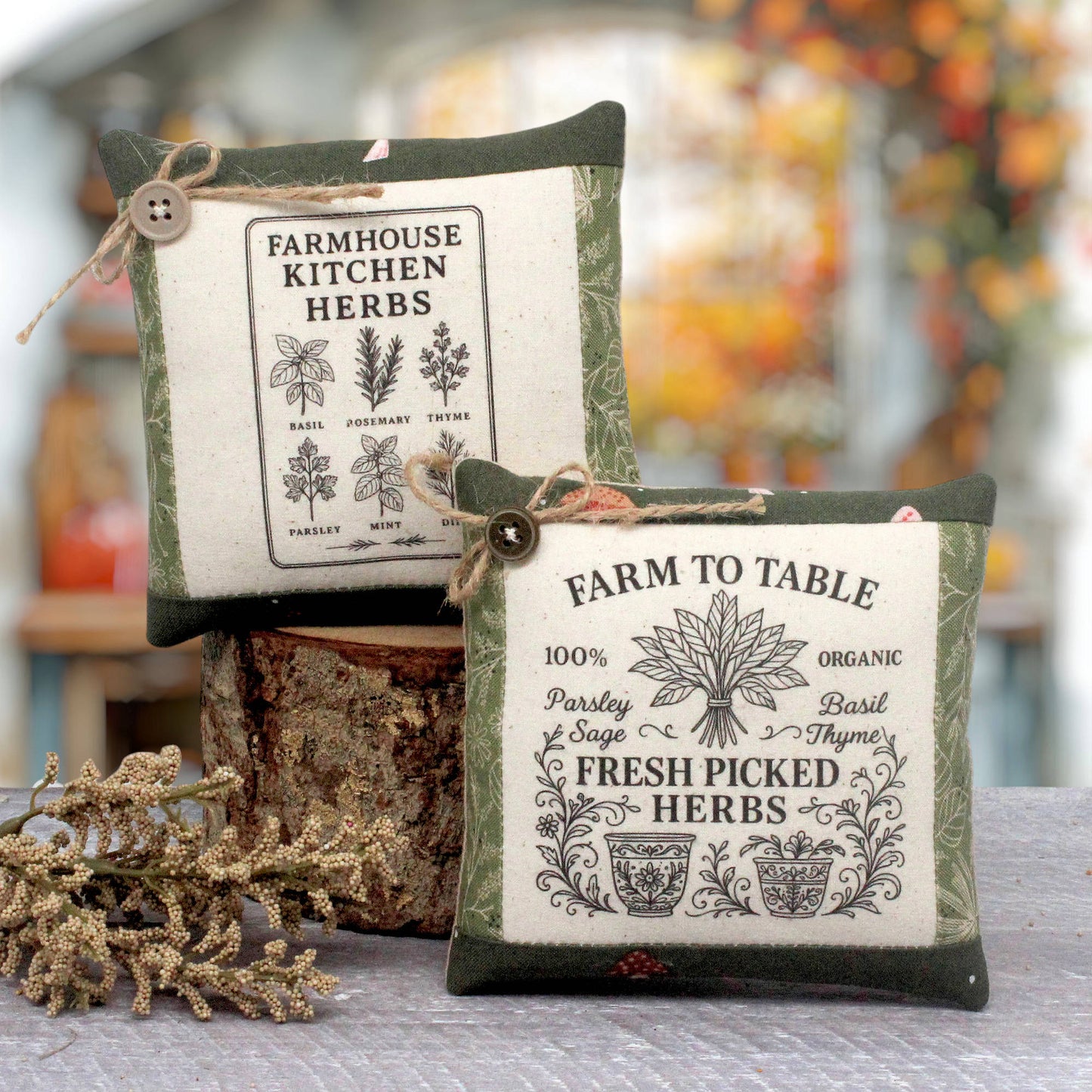 Two decorative fabric herb mini pillows with vintage-style printed labels, displayed on a rustic wood stump with dried botanicals in the foreground. The background is a softly blurred outdoor garden setting.