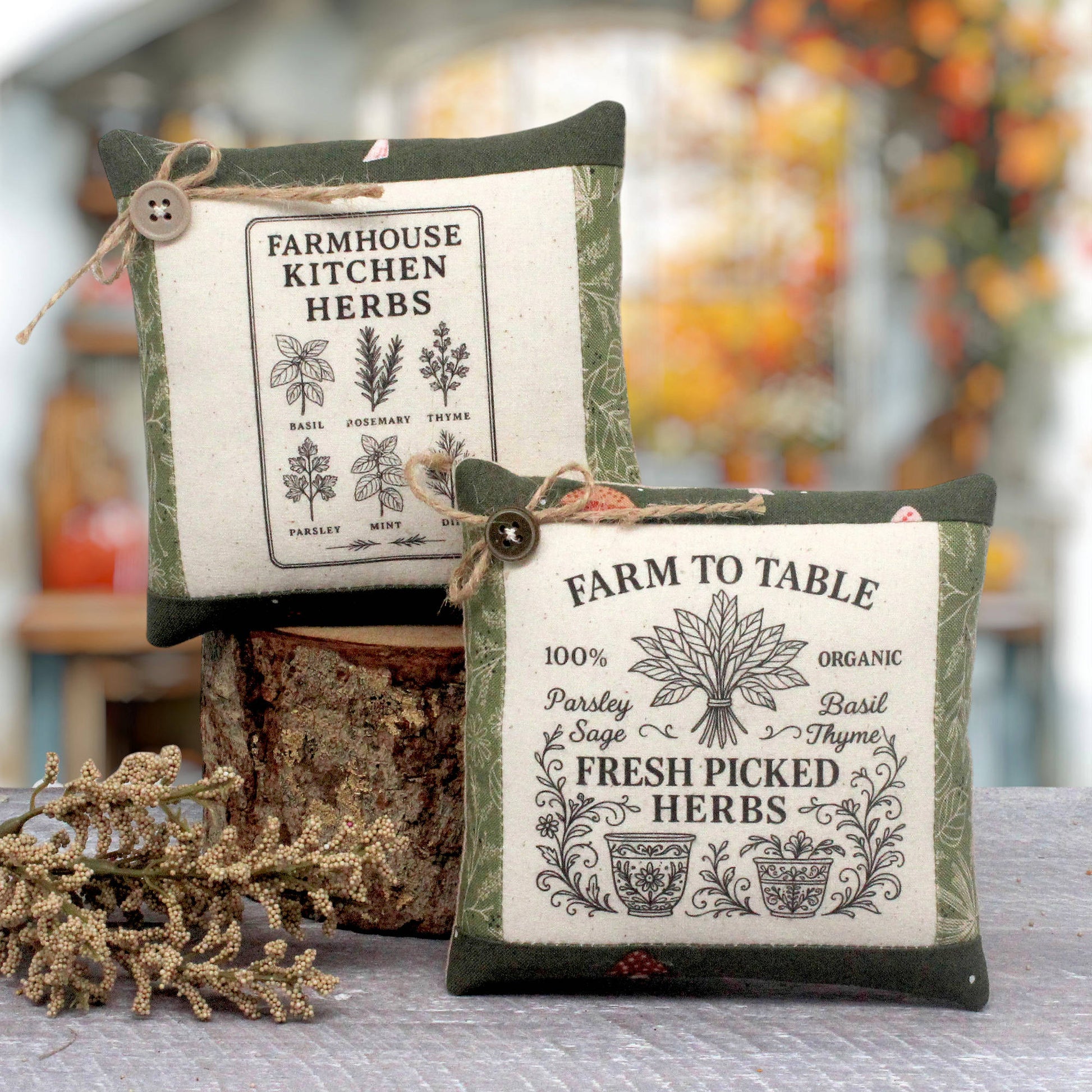 Two decorative fabric herb mini pillows with vintage-style printed labels, displayed on a rustic wood stump with dried botanicals in the foreground. The background is a softly blurred outdoor garden setting.