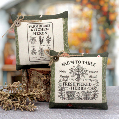 Two decorative fabric herb mini pillows with vintage-style printed labels, displayed on a rustic wood stump with dried botanicals in the foreground. The background is a softly blurred outdoor garden setting.