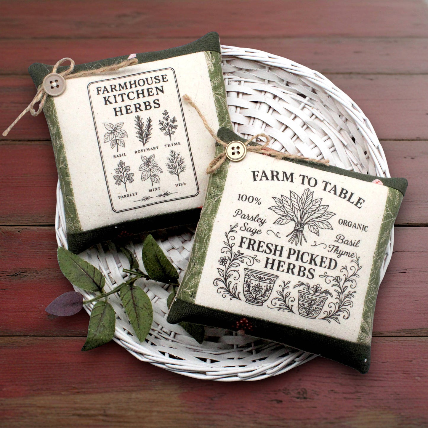 Two small square fabric herb garden mini pillows with vintage-style printed labels, displayed on a white woven tray over a dark red wooden surface.