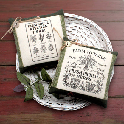 Two small square fabric herb garden mini pillows with vintage-style printed labels, displayed on a white woven tray over a dark red wooden surface.