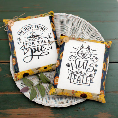 Two decorative fabric pillows displayed on a white wicker tray over a green wooden surface. One pillow reads "I'M JUST HERE FOR THE pie" with a pie illustration, and the other reads "Nuts about FALL" with a squirrel illustration.