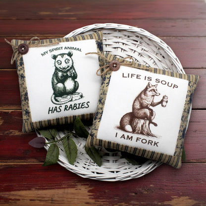 Two decorative square mini pillows displayed on a white wicker tray over a rustic red wooden surface. One pillow shows a black-and-white opossum illustration with the text 'MY SPIRIT ANIMAL HAS RABIES,' and the other shows a brown fox illustration with the text 'LIFE IS SOUP I AM FORK.