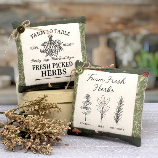 Two decorative fabric herb-themed mini pillows styled on a table with dried herbs. One pillow reads 'Farm to Table 100% Organic Fresh Picked Herbs' with sage, mint, and basil illustrations.
