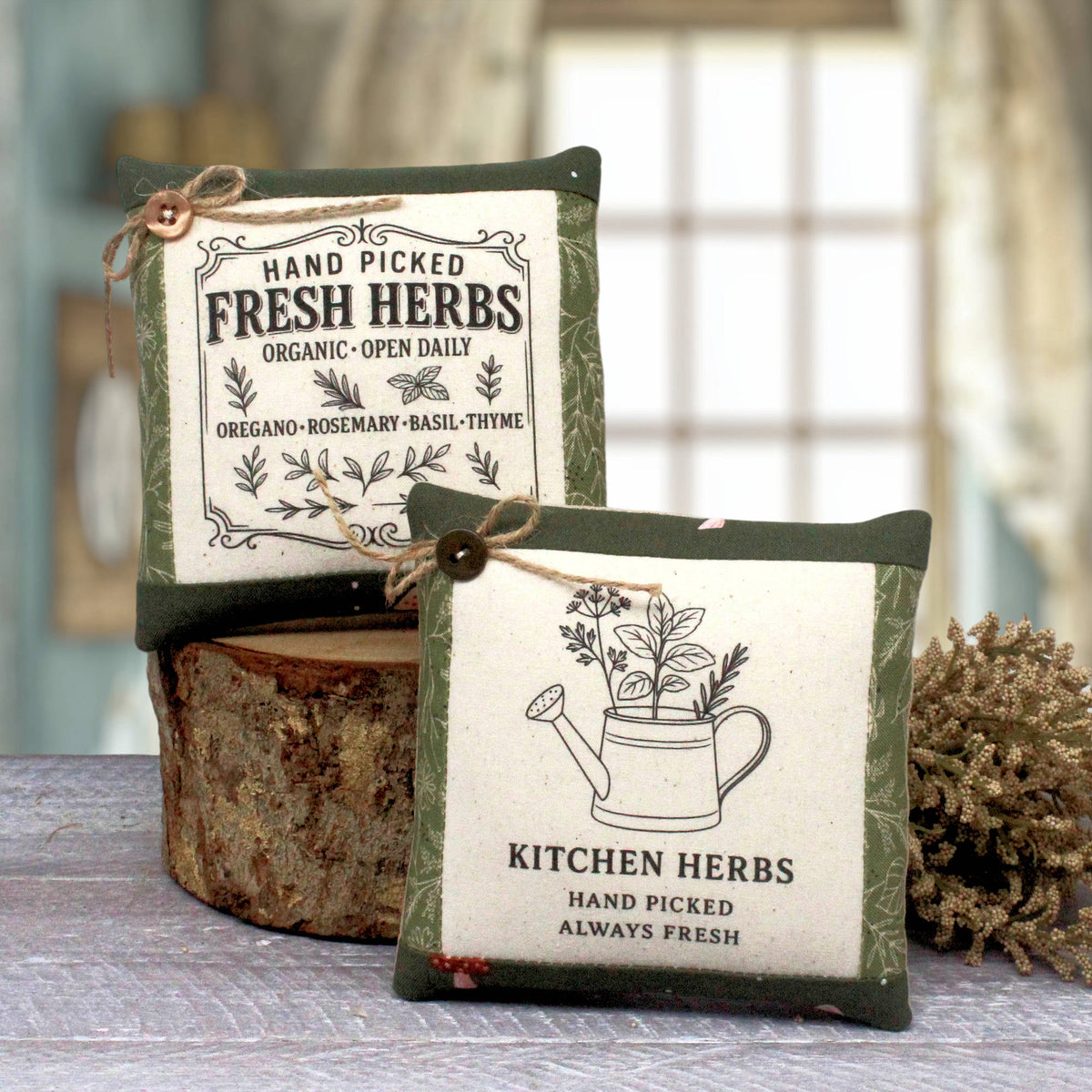 Two decorative square herb mini pillows with green borders and cream centers, tied with twine, displayed on a rustic wood slice with a blurred kitchen background.