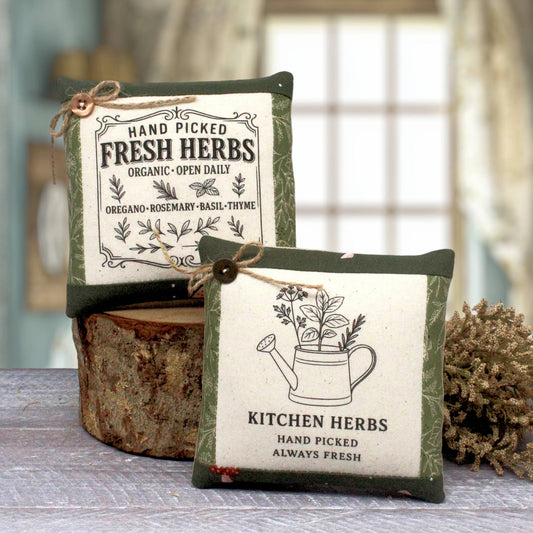 Two decorative square herb mini pillows with green borders and cream centers, tied with twine, displayed on a rustic wood slice with a blurred kitchen background.