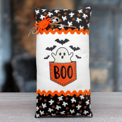 A Halloween-themed fabric pillow or pouch with embroidered ghosts, bats, and the word 'BOO' in orange on a white center panel, with black patterned borders and an orange spider decoration, photographed against a blurred indoor background.