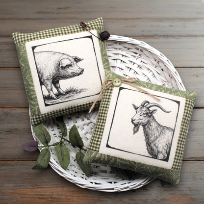 Two decorative square fabric pillows with gingham trim and black-and-white animal illustrations, displayed on a white woven tray over a rustic wooden surface.