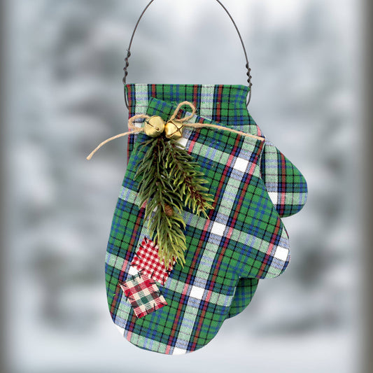 A Christmas ornament shaped like a mitten, made from green, blue, and white plaid fabric, decorated with a sprig of pine, small gold bells, twine, and a jute twine bow, hanging against a blurred gray background.