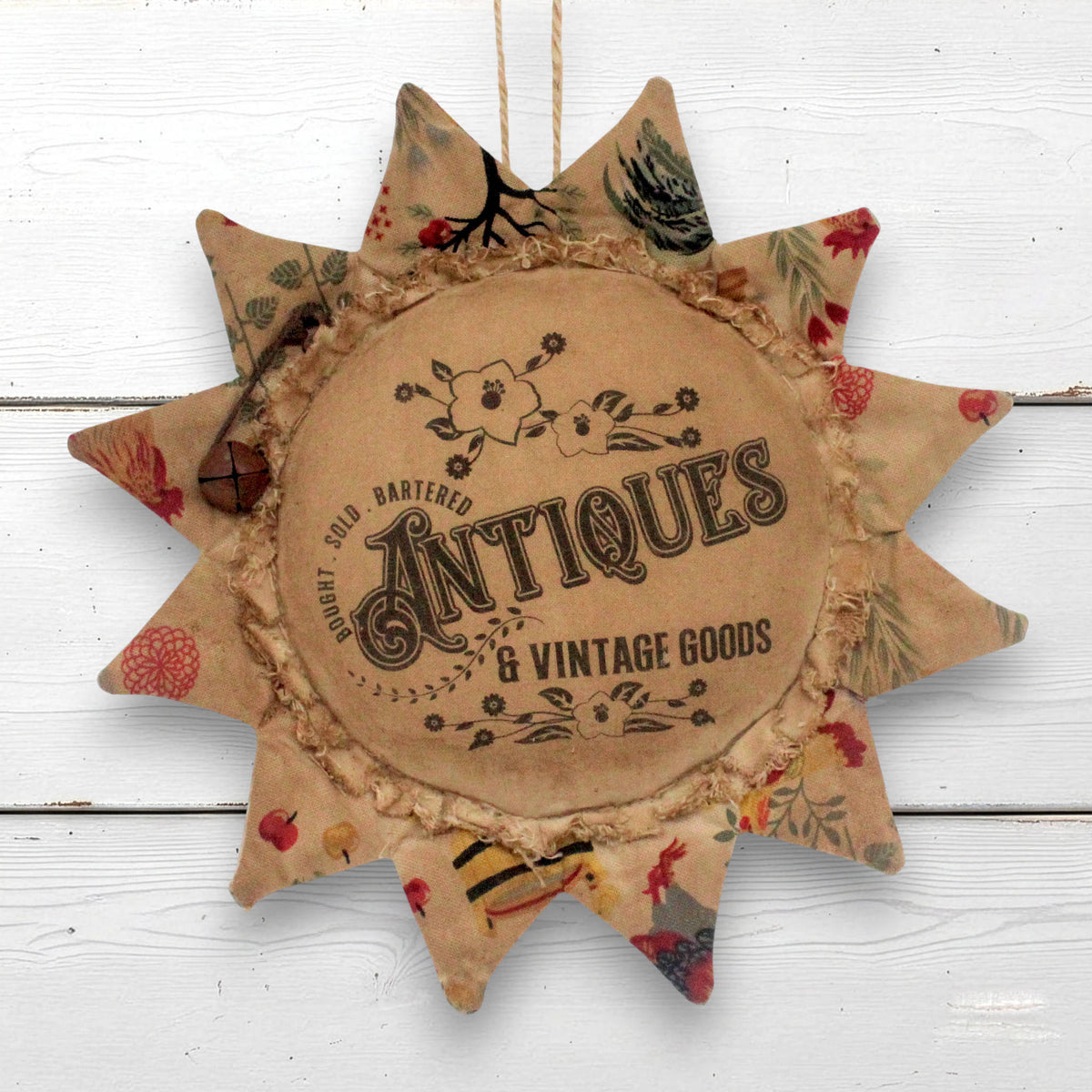 A rustic fabric sunflower-shaped hanging ornament with a central round patch reading 'Antiques & Vintage Goods' and printed vintage-style lettering and floral motifs. The ornament has multicolored patterned points and is shown hanging against a white painted wood background.