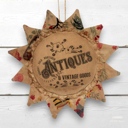 A rustic fabric sunflower-shaped hanging ornament with a central round patch reading 'Antiques & Vintage Goods' and printed vintage-style lettering and floral motifs. The ornament has multicolored patterned points and is shown hanging against a white painted wood background.
