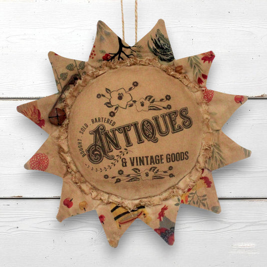 A rustic fabric sunflower-shaped hanging ornament with a central round patch reading 'Antiques & Vintage Goods' and printed vintage-style lettering and floral motifs. The ornament has multicolored patterned points and is shown hanging against a white painted wood background.
