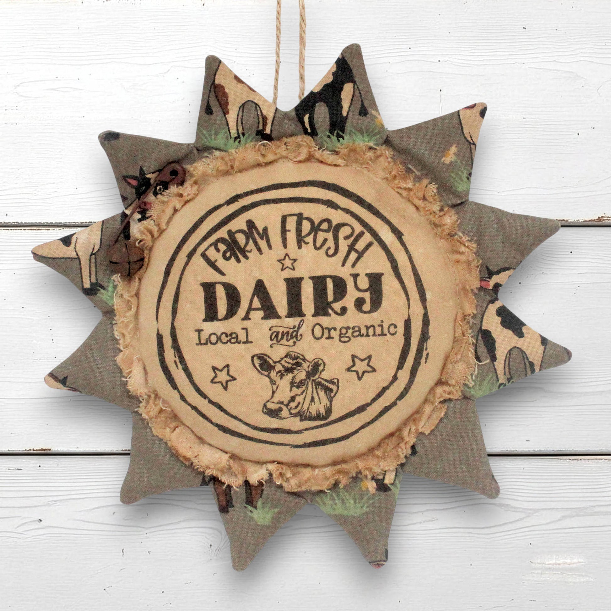 A rustic fabric sunflower-shaped hanging ornament with a central round sign reading 'Farm Fresh Dairy Local and Organic' and a cow illustration, shown against a white wood plank background.