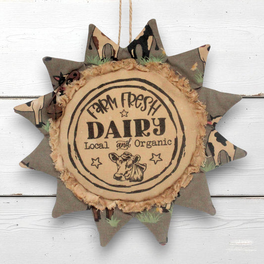 A rustic fabric sunflower-shaped hanging ornament with a central round sign reading 'Farm Fresh Dairy Local and Organic' and a cow illustration, shown against a white wood plank background.