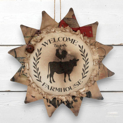 A rustic sunflower-shaped hanging wall decor sign with a central circular panel reading “WELCOME to our FARMHOUSE” and a black cow-and-rooster silhouette, surrounded by distressed beige, brown, and red patchwork star points.