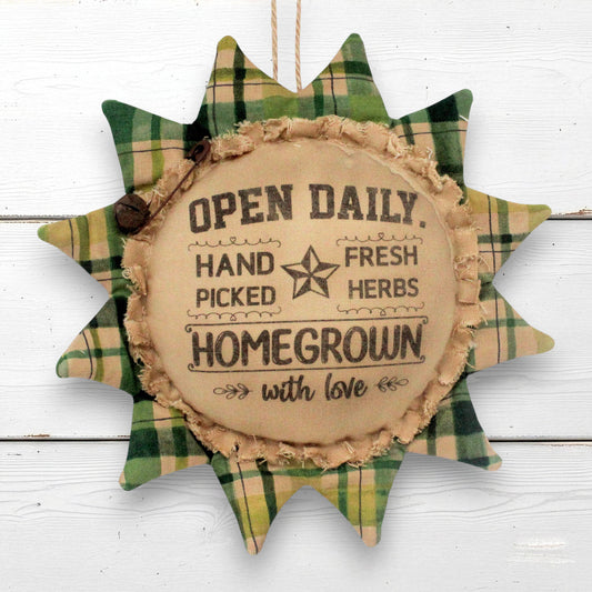 A rustic sunflower fabric hanging ornament with a plaid green and tan border and a tan center that reads, 'OPEN DAILY. HAND PICKED FRESH HERBS HOMEGROWN with love.