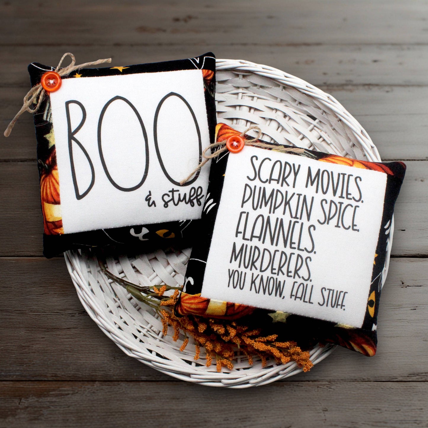 Two Halloween-themed fabric mini pillows displayed on a white wicker tray over a rustic wood surface. One square white pillow reads “BOO in stuff” with black lettering and a small orange button; the other reads “SCARY MOVIES, PUMPKIN SPICE, FLANNELS, MURDERERS, YOU KNOW, FALL STUFF” with black lettering and a small orange button.