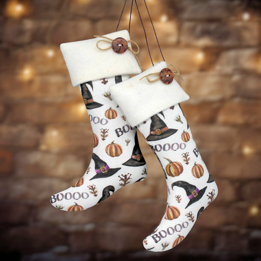 Two white Christmas stockings with a witch and pumpkin print, hanging against a warm, blurred brick wall background with bokeh lights.