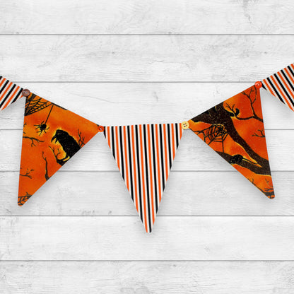 Halloween bunting banner with triangular flags featuring orange spiderwebs, black crows, and black-and-orange vertical stripes, hung against a white wood plank background.