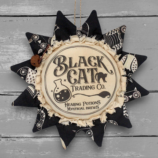 A handmade sunflower ornament for Halloween, featuring the text 'Black Cat Trading Co' and 'Healing Potions Mystical Brews' on a cream center, surrounded by black patterned fabric petals, hanging against a gray wood plank background.