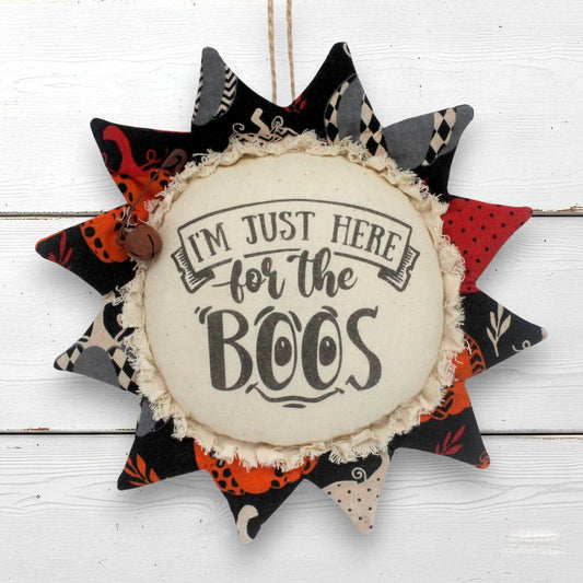 A fabric sunflower hanging ornament with a rustic twine loop and a central cream-colored circle that reads “I’M JUST HERE for the BOOS.” The surrounding starburst points are made from patterned fabrics in black, orange, red, gray, and beige, arranged on a white wood plank background.