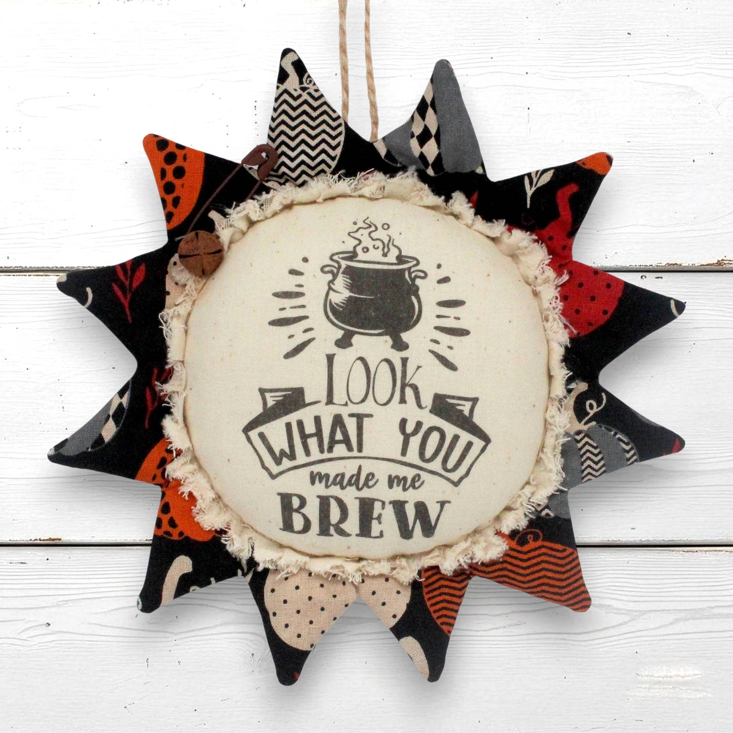 a primitive sunflower fabric ornament for halloween with a center that reads "Look what you made me brew" decorating with a rusty safety pin and bell