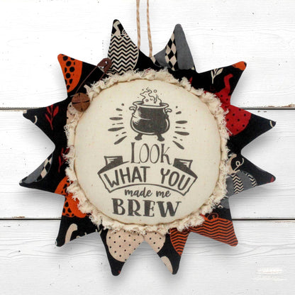 a primitive sunflower fabric ornament for halloween with a center that reads "Look what you made me brew" decorating with a rusty safety pin and bell