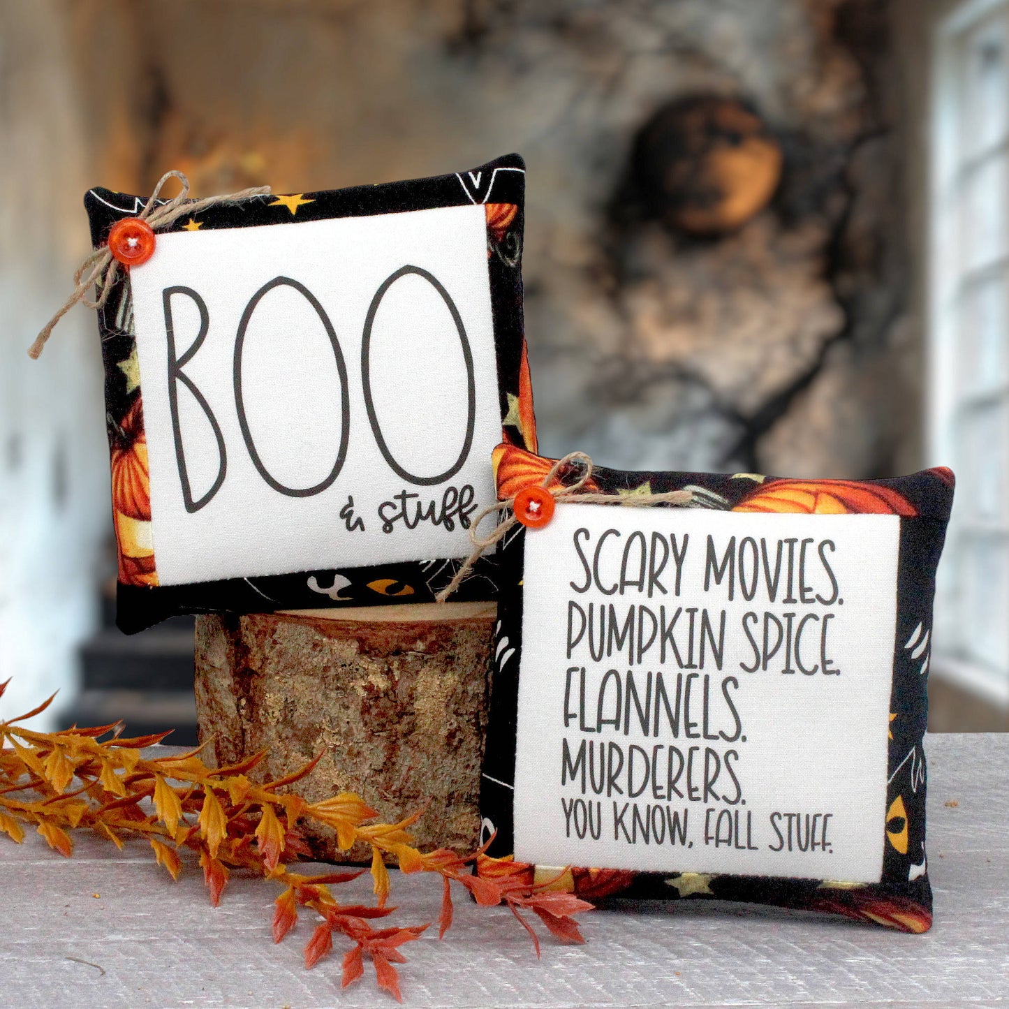 Two Halloween-themed fabric mini pillows displayed on a white wicker tray over a rustic wood surface. One square white pillow reads “BOO in stuff” with black lettering and a small orange button; the other reads “SCARY MOVIES, PUMPKIN SPICE, FLANNELS, MURDERERS, YOU KNOW, FALL STUFF” with black lettering and a small orange button.
