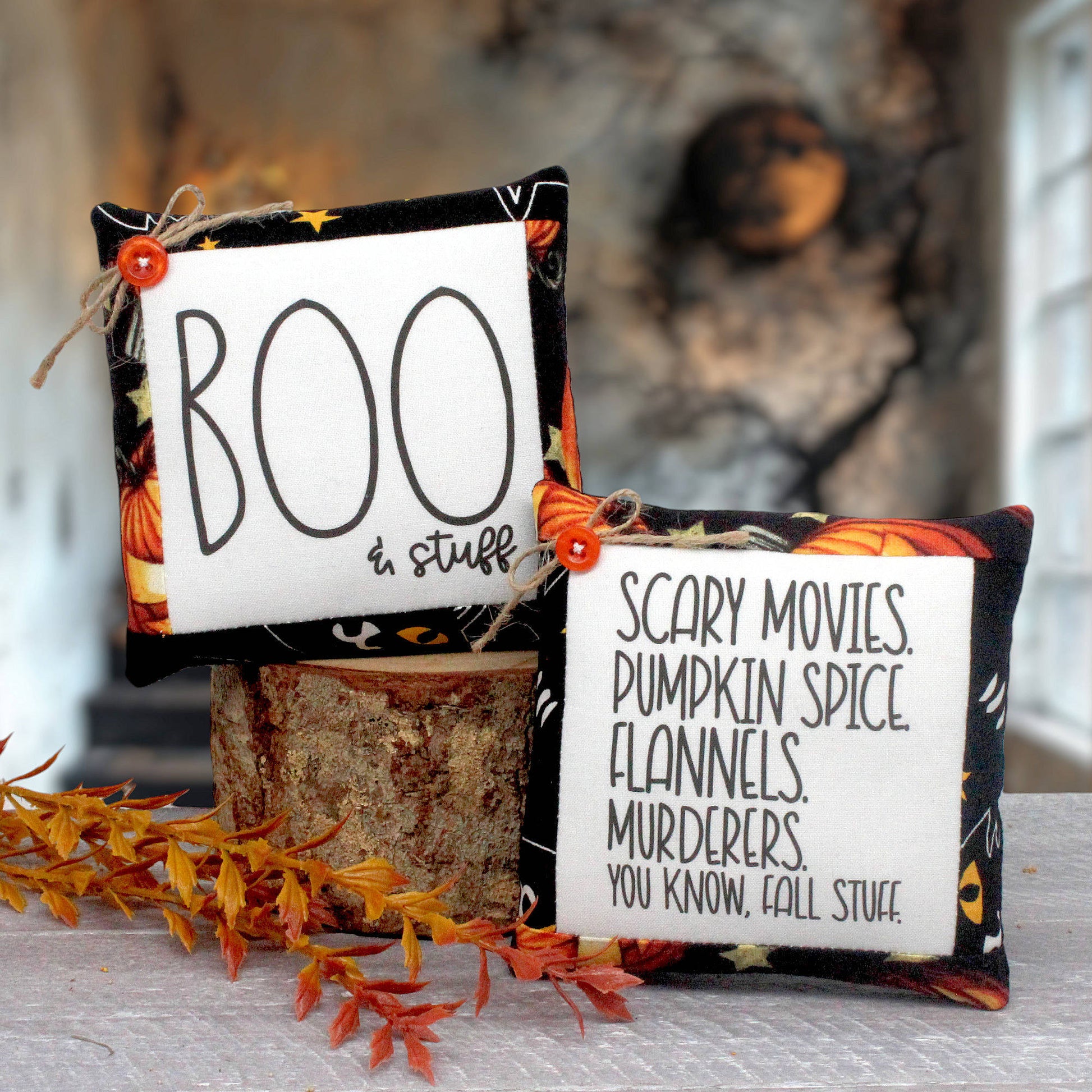 Two Halloween-themed fabric mini pillows displayed on a white wicker tray over a rustic wood surface. One square white pillow reads “BOO in stuff” with black lettering and a small orange button; the other reads “SCARY MOVIES, PUMPKIN SPICE, FLANNELS, MURDERERS, YOU KNOW, FALL STUFF” with black lettering and a small orange button.