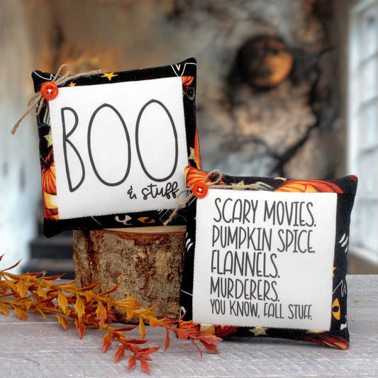 Two Halloween-themed fabric mini pillows displayed on a white wicker tray over a rustic wood surface. One square white pillow reads “BOO in stuff” with black lettering and a small orange button; the other reads “SCARY MOVIES, PUMPKIN SPICE, FLANNELS, MURDERERS, YOU KNOW, FALL STUFF” with black lettering and a small orange button.